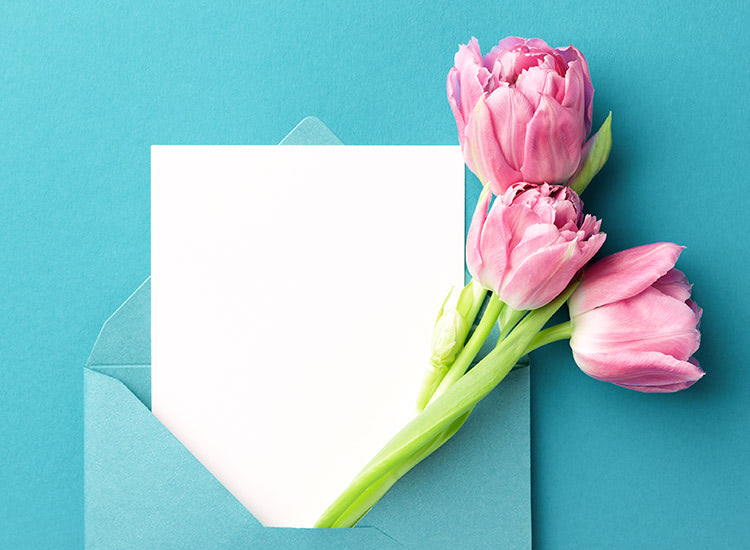 A turquoise envelope holds a white card and three pink tulips, set against a matching turquoise background.