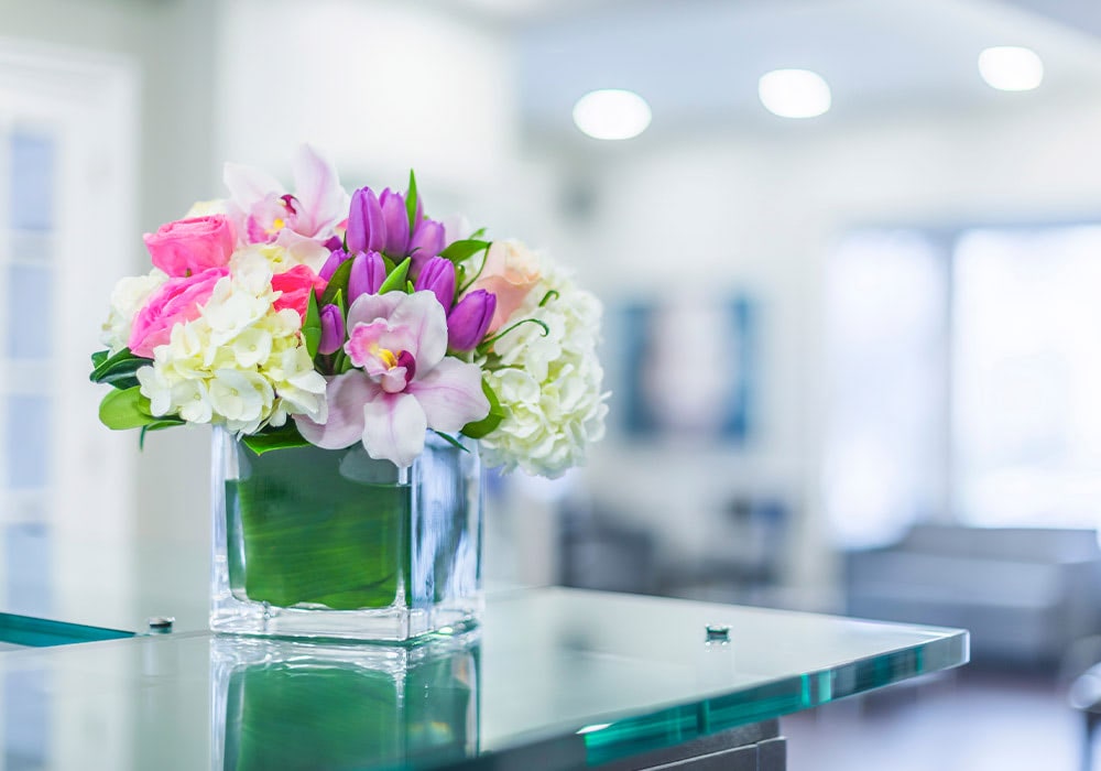 A vase of vibrant mixed flowers, including pink roses and white hydrangeas, sits on a glossy glass table in a brightly lit, modern interior space.