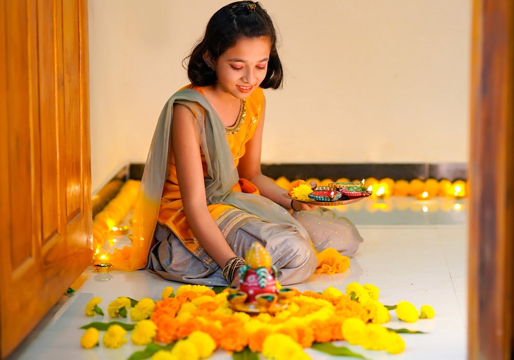 A woman in traditional attire, sitting and holding a decorated plate while arranging marigold flowers and festive items on the floor, is surrounded by lights and more flowers.