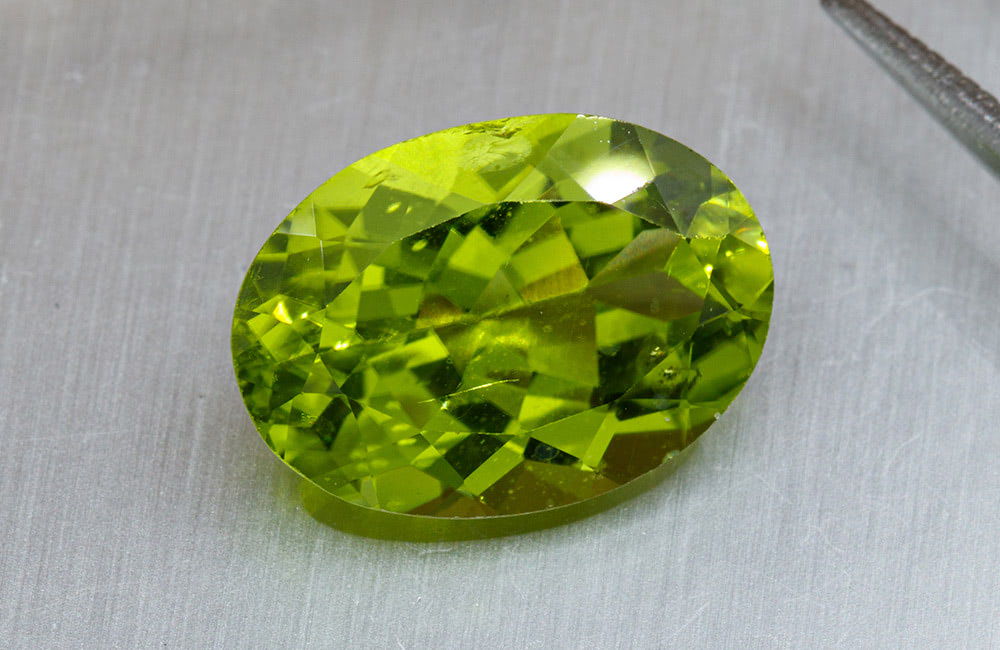 An oval-cut, faceted yellow-green gemstone rests on a smooth, silvery-gray surface with a metal object nearby.