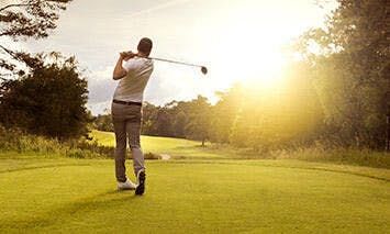 A golfer swings a club on a well-manicured course, with trees surrounding the area and the sun brightly shining in the background.