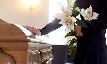 A person in black attire holds a white lily bouquet and rests a hand on a wooden casket, set in a softly lit room.