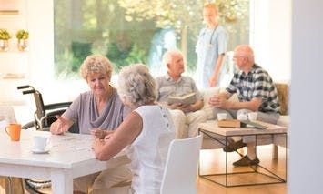 Elderly women sitting at a table working on a puzzle, while a nurse and two elderly men interact in a bright, modern living room. An empty wheelchair is nearby.