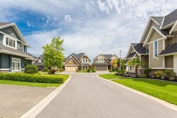 A row of modern suburban houses line a wide, clean street under a partly cloudy sky; the well-kept lawns and trees enhance the picturesque neighborhood ambiance.